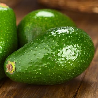 Avocado 'Fuerte' fruit with glossy, smooth green skin and small yellow speckles, resting with two more avocados on a wooden surface.