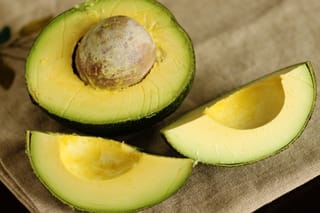 Avocado 'Reed' fruit cut open, showing pale yellow-green flesh and a round brown seed, with two wedges on a linen cloth.