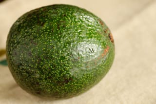 Avocado 'Reed' fruit, round with dark green pebbly skin covered in small yellow speckles and a few brown marks, resting on cream fabric.