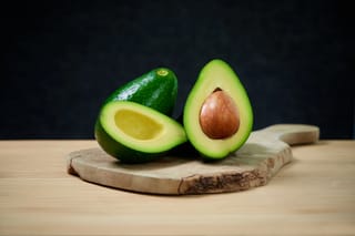 Avocado 'Zutano' fruit halved on a rustic wooden board, showing pale green flesh, a brown seed and glossy dark green skin.