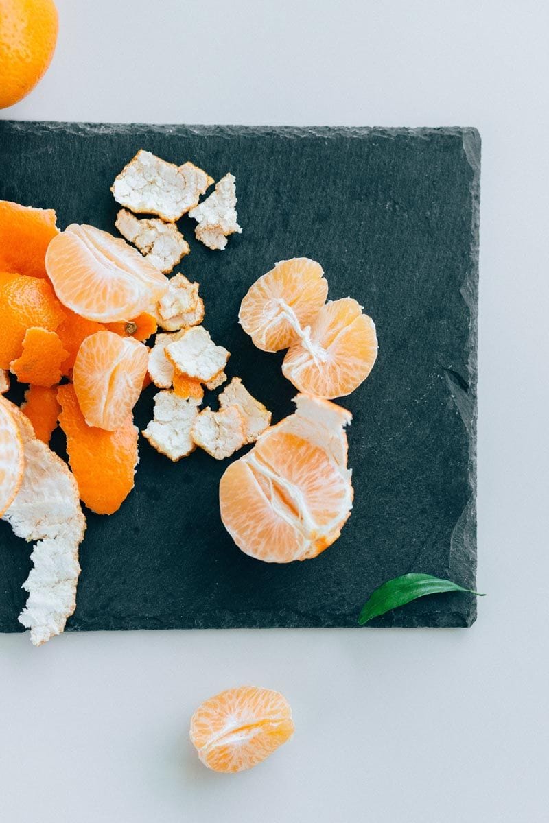 Peeled Citrus mandarin 'Okitsu' (mandarin) segments and orange peel pieces on a dark slate board, with a single green leaf, seen from above.