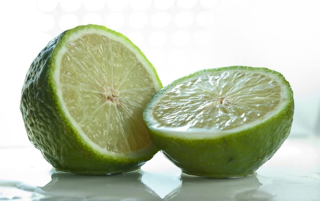 Citrus lime 'Bearss' (Persian Lime) fruit cut in half, showing pale green juicy flesh and dimpled green skin on a glossy white surface.