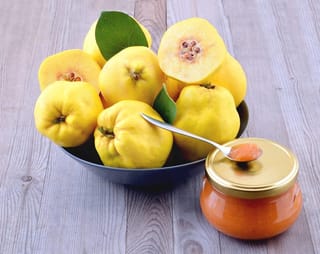 Quince 'Taihape' (Cydonia oblonga) fruit, yellow and whole or halved, in a dark bowl beside a jar of orange quince preserve with a spoon.