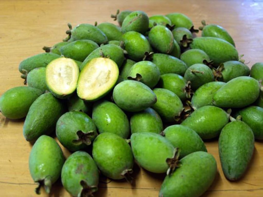 Feijoa sellowiana 'Golden Goose' (feijoa) fruit, a pile of green oval fruit on a wooden surface, with one fruit halved to show pale flesh.