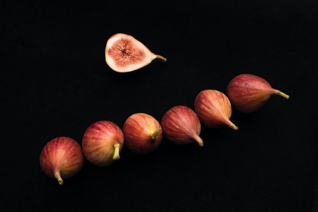 Ficus carica 'Brown Turkey' fig fruit: six whole purplish-brown figs with green stalks in a row beside a halved fig showing pink flesh, on black.