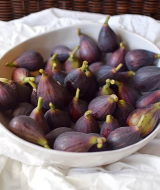 Harvested Ficus carica 'Brunoro Black' figs with purple-black skin and green stalks, heaped in a white bowl on crumpled white cloth.