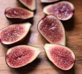 Halved Ficus carica 'Brunoro Black' fig fruit on a wooden board, showing dark purple skin, pale rind and red seeded flesh.