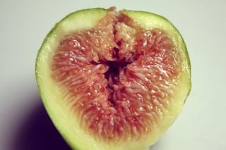 Halved Ficus carica 'Lesa' fig fruit showing pink-red flesh with small seeds inside a pale green-yellow skin, on a plain white background.