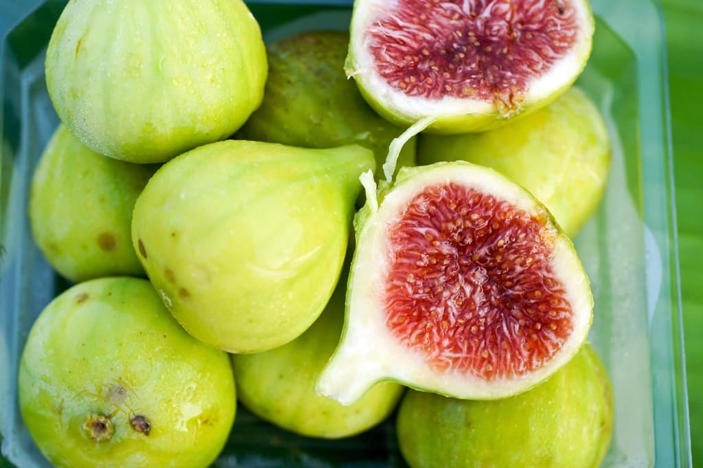 Ficus carica 'Lesa' fig fruit with pale green-yellow skin in a clear tray, two halved to show red-pink, seed-filled flesh.
