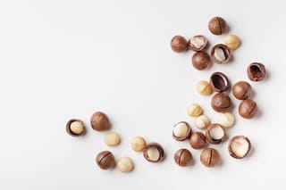 Macadamia 'GT207' nuts scattered on a white surface, with brown round shells, some cracked open to show the cream kernels.