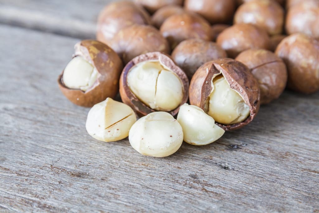 Macadamia 'Beaumont' nuts on a weathered wooden surface, with brown shells cracked open to show the creamy white kernels.