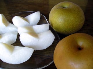 Pear 'Hosui' fruit: two round Asian pears with speckled golden-brown skin beside a glass plate of peeled white slices on a wooden table.