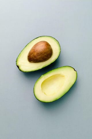 Avocado 'Bacon' (Persea americana) fruit halved to show pale yellow-green flesh and a brown stone, on a plain grey-blue background.
