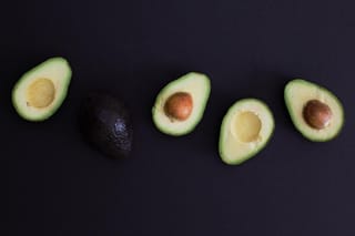 Avocado 'Hass' fruit on a dark background: four halves showing pale green flesh and brown stones, beside one whole pebbly, purple-black fruit.