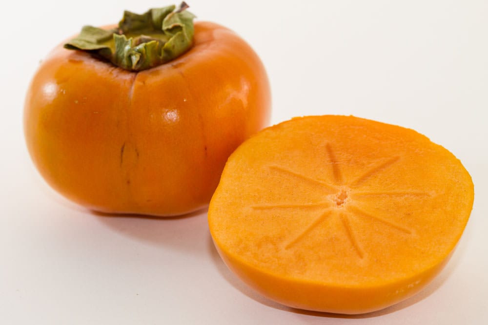 Persimmon 'Fuyu' (Japanese Persimmon) fruit: one whole orange fruit with a dried green calyx beside a halved fruit showing smooth flesh, on white.
