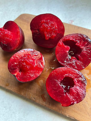 Plum 'Sultan' fruit halved on a wooden board, showing glossy deep-red skin and juicy red flesh with stone cavities.