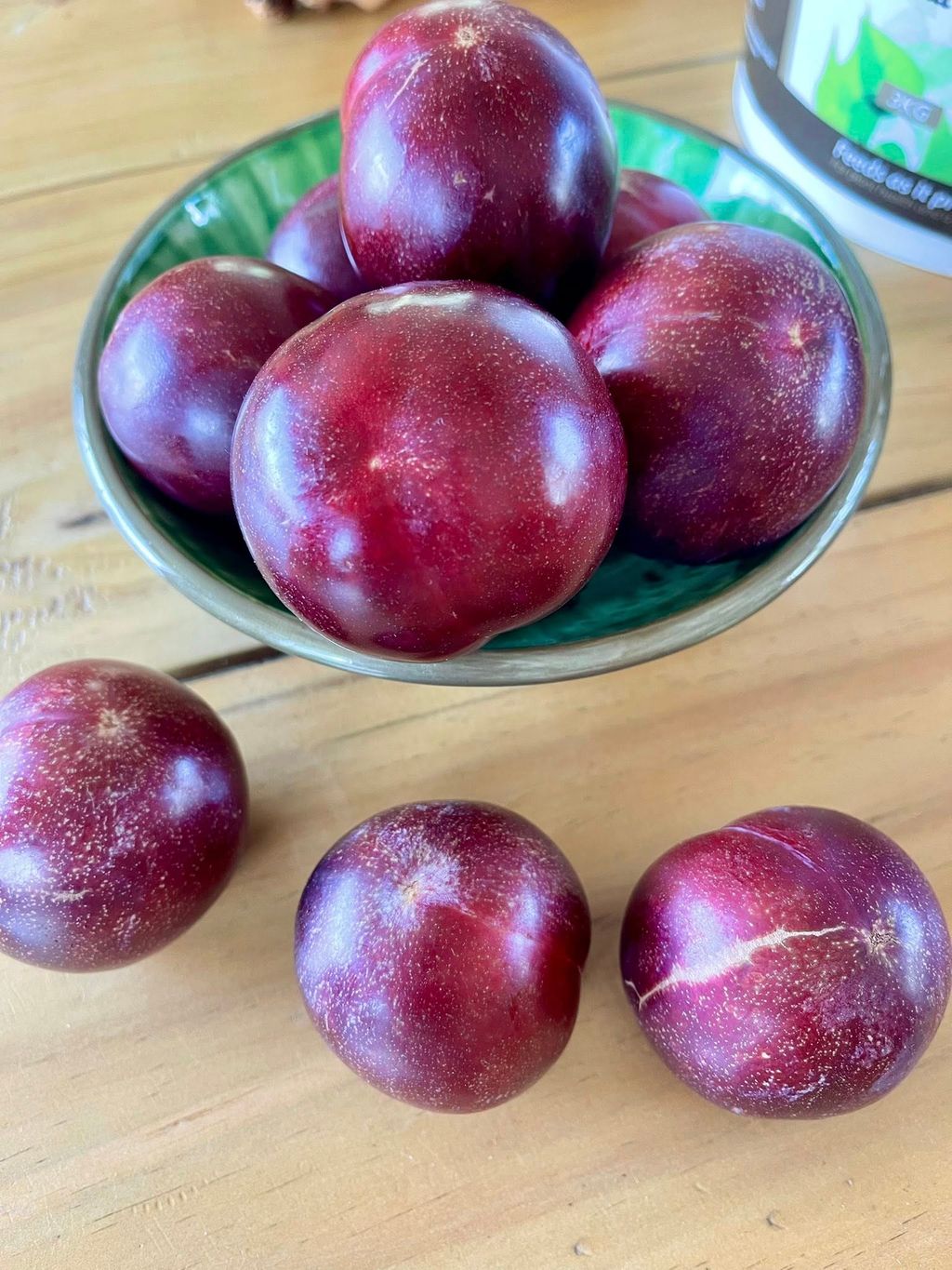 Plum 'Sultan' fruit with glossy deep-red skin, heaped in a green ceramic bowl with three more plums on a wooden table.