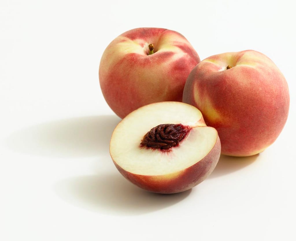 Peach 'April White' fruit: two whole peaches with a pink-red blush over cream-yellow skin beside a halved fruit showing white flesh and a dark stone.