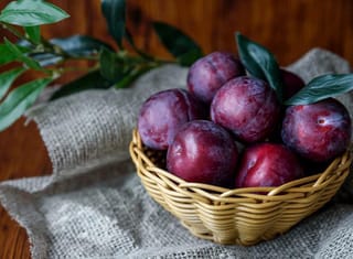 Plum 'Billington' (Japanese Plum) fruit with deep red-purple skin heaped in a woven basket on hessian, beside green leafy stems.