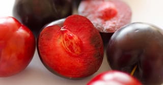 Plum 'Billington' (Japanese Plum) fruit, whole and halved, showing dark red-purple skin, bright red flesh and a stone on a pale surface.