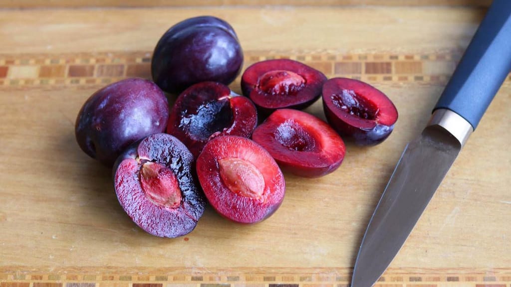 Plum 'Black Doris' (Japanese Plum) fruit, whole and halved to show deep red flesh and stones, on a wooden board beside a knife.