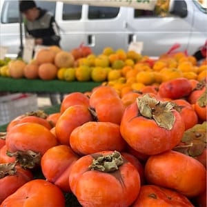 Persimmon 'Fuyu' (Diospyros kaki, Japanese Persimmon): piles of squat orange fruit with green-brown calyxes on a market stall, with more fruit and a vehicle behind.