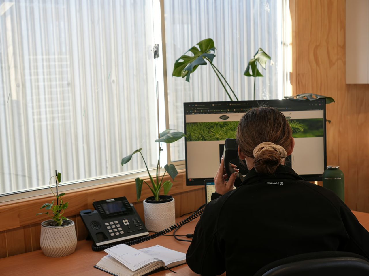 Woman in black jacket on phone at bright office desk with computer, phone, open notebook, and potted plants. Large window with blinds and wood paneling in background.