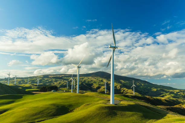 Wind turbines stand tall on rolling green hills under a bright blue sky dotted with fluffy white clouds. A sustainable energy landscape with wind power generation.