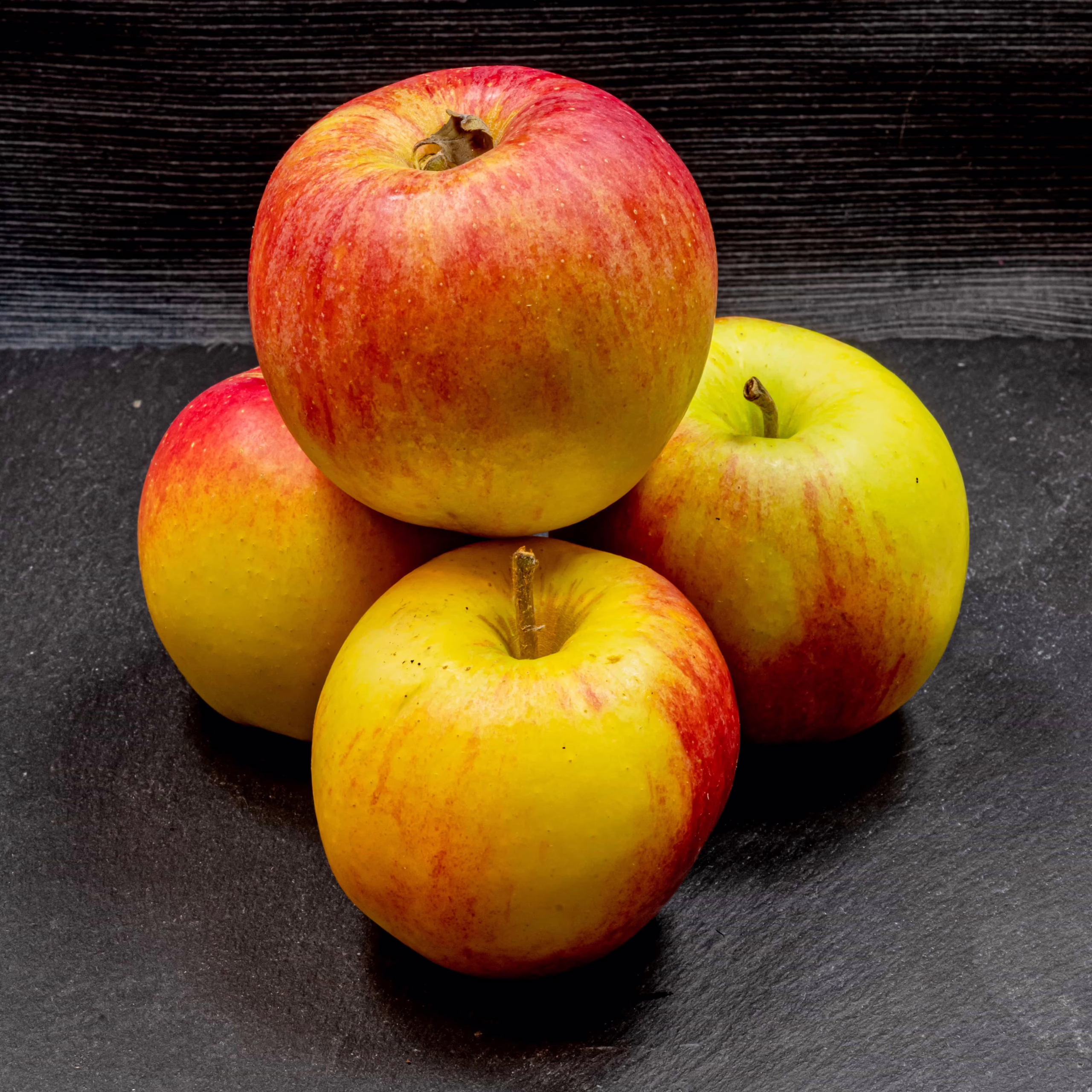 Pile of four 'Initial' apples. Yellow and red skin, round shape, with stems visible on some.