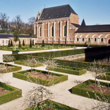 Formal garden with Buxus sempervirens hedges, grid pattern, leading to brick building with arched windows.