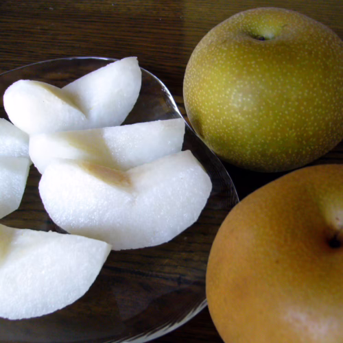 Pear ‘Hosui’: Two whole, speckled golden-brown Asian pears plus slices on a clear plate, showcasing juicy, white flesh.