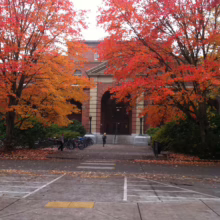 Autumn at Oregon State University: Vibrant red and orange trees frame the archway entrance to a brick building, with students walking by bicycles in the foreground.