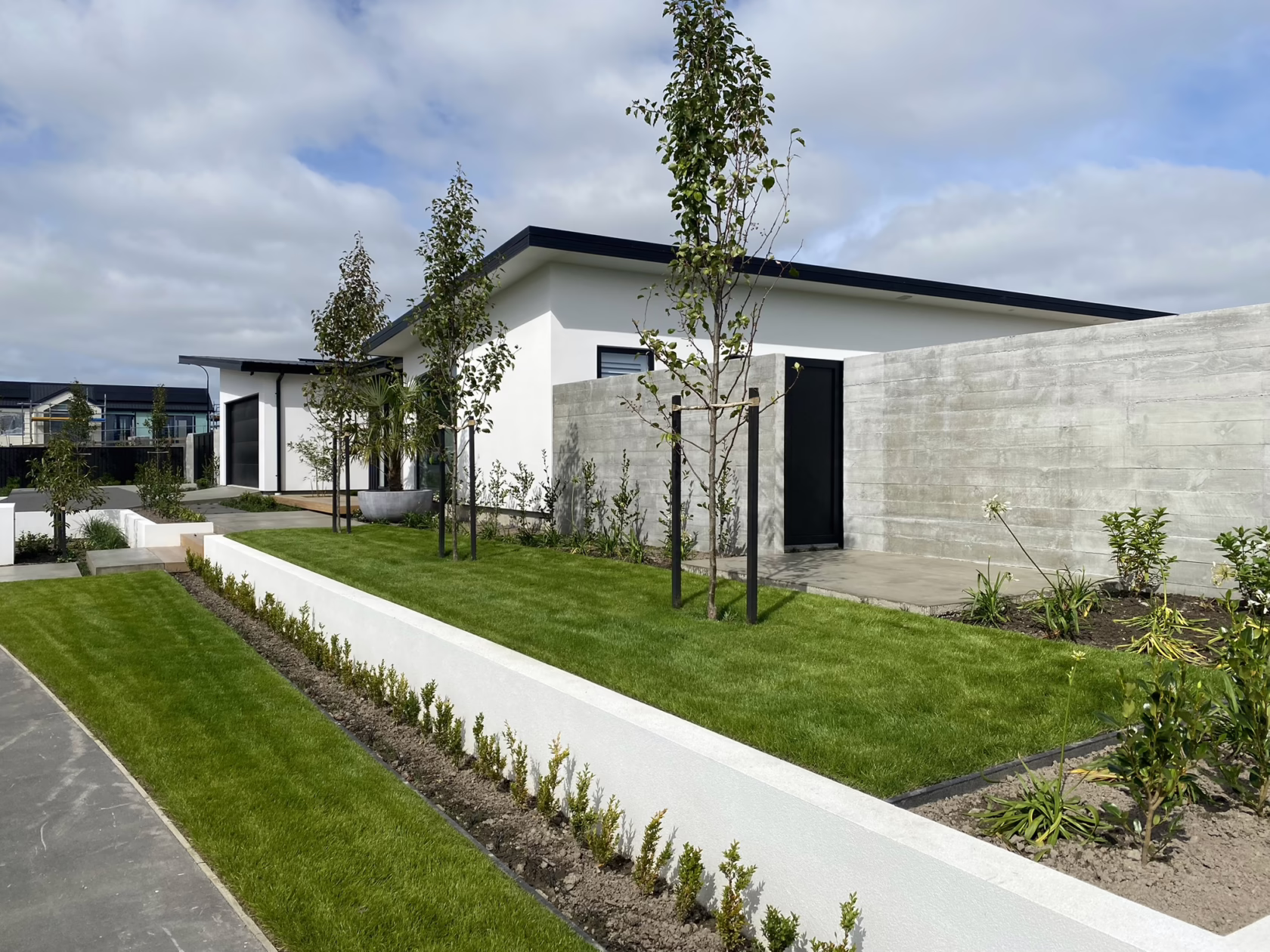 Modern landscape design featuring Pyrus calleryana 'Slim Jim' trees in a neatly manicured lawn, bordered by white walls and a concrete fence, against a white house under a cloudy sky.
