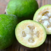 Feijoa sellowiana 'Bambina': Green, egg-shaped fruit, one whole, one halved, revealing juicy pulp and seeds on a rustic wooden surface.