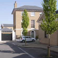 Three-story yellow brick house with black iron fence and white car parked in driveway. Two young trees flank the scene, adding a touch of green to the urban landscape.