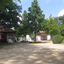 Park scene with trees shading a sandy courtyard, benches, and buildings with red roofs under a blue sky. Green foliage and flowers add color to the serene outdoor setting.