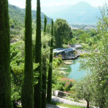 Lush garden scene with tall, slender cypress trees framing a tranquil pond. Water lilies float on the pond's surface, with a cafe and mountains visible in the background.