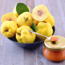 Bowl of bright yellow quince fruit, some cut open to reveal seeds, with a jar of quince jam and a spoonful on the lid, set on a rustic wooden table.