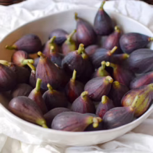 Bowl of ripe, dark purple figs with green stems, sitting on a white cloth. The figs are freshly picked and ready to eat.