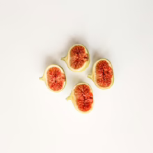 Four halved figs, their red pulps facing up, arranged in a square on a white surface. The figs have light green skins and are ripe and juicy.