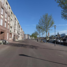 Brick buildings line a wide brick walkway along a canal in a Dutch city. Trees, lampposts, and boats dot the waterway under a clear blue sky, capturing the serene urban landscape.