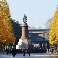 Statue of Prince Arisugawa Taruhito in Tokyo's Ueno Park, surrounded by vibrant yellow ginkgo trees in autumn. Pedestrians stroll along the pathway toward the torii gate of Toshogu Shrine.