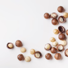 Scattered macadamia nuts, some shelled, on a clean white surface. The nuts range from creamy white kernels to brown, hard shells.