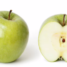 A whole, shiny green apple sits next to a halved apple, revealing its light flesh and dark seeds. The halved apple shows the stem and core, highlighting the fruit's natural texture.