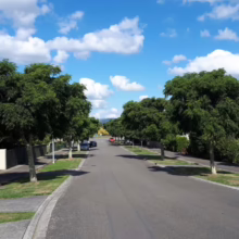 Tree-lined suburban street under a bright blue sky dotted with puffy white clouds. A peaceful residential scene with neat lawns and parked cars.