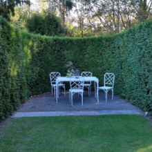 White wrought iron table and chairs nestled in a secluded garden nook surrounded by tall, curved hedges. A peaceful outdoor dining space.
