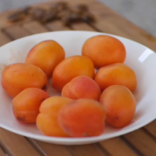 A white plate filled with vibrant orange apricots on a wooden table. Some apricots are also scattered in the background.