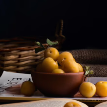Bowl of ripe yellow plums with leaves, sitting on a book. A rustic basket and burlap fabric are in the background, creating a warm, natural still life.