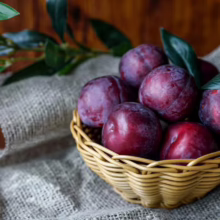 A small woven basket overflowing with ripe, deep purple plums, nestled on a burlap cloth with green leaves in the background. Fresh, juicy plums ready to eat.