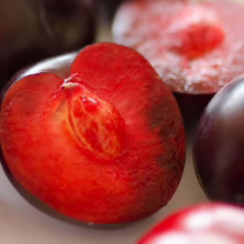 Close-up of ripe plums, some whole with deep purple skin, one cut open revealing juicy red flesh and a pit. The plums are on a white surface.
